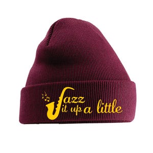 Può includere: Un berretto bordeaux con risvolto. Il berretto presenta la scritta "Jazz it up a little" in giallo, insieme a una grafica di sassofono e note musicali. Il berretto è realizzato in maglia a coste.