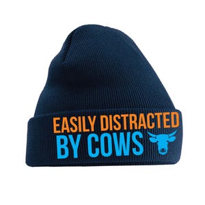 May include: A navy blue knitted beanie hat with the slogan "EASILY DISTRACTED BY COWS" in orange and blue. A blue cow graphic is on the right side of the folded brim. A winter hat.