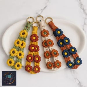 May include: Four handmade macrame keychains with floral designs. Each keychain features a different color scheme: green and yellow, yellow and red, cream and orange, and orange and blue. Gold-colored key rings are attached to the top of each keychain. The bottom left corner has a logo with the text "LXTEEN ARTSHOP".