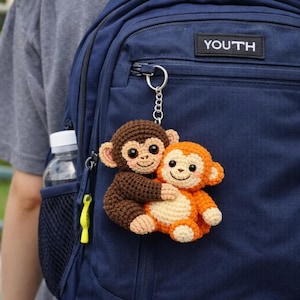May include: A handmade crochet keychain featuring two hugging monkeys. One monkey is brown, and the other is orange. The keychain is attached to a navy blue backpack. The word "YOUTH" is visible on the backpack.