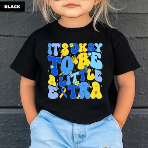 May include: Black t-shirt with the text "IT'S OKAY TO BE A LITTLE EXTRA" in blue and yellow. The design includes flowers and ribbons. The child is wearing blue jeans.