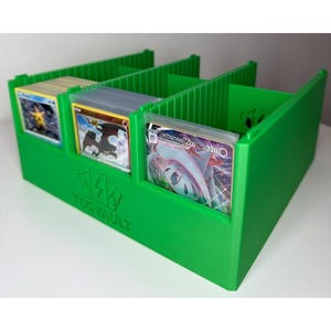 Può includere: Un organizer per carte verde brillante con tre scomparti, contenente varie carte collezionabili. L'organizer ha il testo "TCGVAULT" sul davanti. Le carte presentano illustrazioni colorate e sono in custodie protettive.