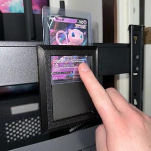 Puede incluir: Un expositor negro que contiene dos cartas de Pokémon. La carta superior presenta un personaje Mew rosa con el texto "Mew V" y "180". La carta inferior es parcialmente visible, con detalles morados y blancos. El expositor está sobre una estantería metálica negra.