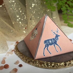 Può includere: Una scatola regalo rosa a forma di piramide con un'illustrazione di cervo blu e un disegno di lanterna bianca. La scatola è su un piatto decorativo grigio e oro con bordo dorato. Cristalli decorativi sparsi.