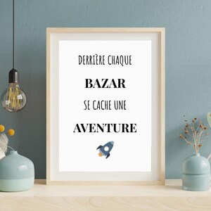 Puede incluir: Impresión enmarcada con el texto "DERRIÈRE CHAQUE BAZAR SE CACHE UNE AVENTURE" y una ilustración de un cohete. El marco es marrón claro y el fondo es blanco. La impresión se muestra en un estante blanco contra una pared azul.