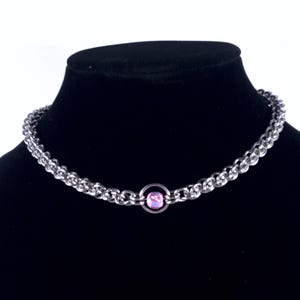 Puede incluir: Un collar de cadena plateada con una gema rosa en el centro. El collar se exhibe en un maniquí negro. La cadena está hecha de pequeños eslabones interconectados. La gema es redonda y está engastada en un anillo plateado.