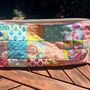 May include: A quilted cosmetic bag with a colorful patchwork design. The bag features a gold zipper and is made of various patterned fabrics, including floral, polka dot, and ladybug prints. The bag is rectangular and sits on a wooden surface.
