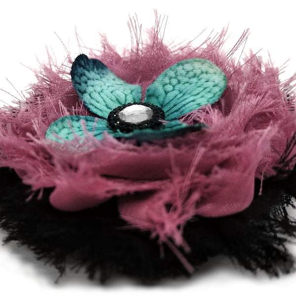 Rosette rose poussiéreuse en détresse surmontée d’une fleur d’aigue-marine bleu-sarcelle et d’un bijou à facettes claires sur un lit de mousseline de soie noire effilochée