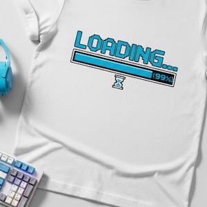 Może przedstawiać: Biała koszulka z niebieską pikselową grafiką "LOADING...", pokazującą 99% postępu. Obraz zawiera również klawiaturę, słuchawki, kontroler do gier i kabel USB, w odcieniach niebieskiego i czarnego.