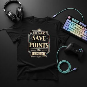 Könnte beinhalten: Schwarzes T-Shirt mit dem Text "LIFE HAS NO SAVE POINTS I DO GAME ON" in einem dekorativen Design. Das Bild enthält auch eine Tastatur, einen Controller und Kopfhörer, alles auf einer schwarzen Oberfläche.