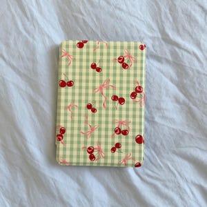 Puede incluir: Funda rectangular para teléfono con un estampado de cuadros vichy verde claro, cerezas rojas y lazos rosas. La funda tiene un acabado brillante y bordes redondeados. El diseño se repite en toda la superficie.