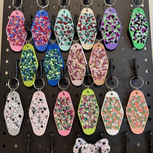 May include: A collection of seventeen diamond-shaped motel-style keychains and one bow-shaped keychain, each decorated with various colored rhinestones. The keychains are hung on a black pegboard display using silver metal split rings.