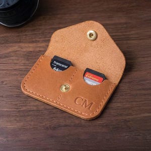 May include: A tan leather SD card holder, open to reveal two memory cards. The holder is secured with a snap closure and features the initials "CM" embossed on the front. The interior is a lighter shade of tan.