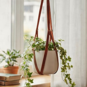 Può includere: Un vaso sospeso con un contenitore in ceramica bianca e cinghie in pelle marrone. Il vaso è sospeso a un anello di metallo e riempito di edera verde. Il portapiante è esposto davanti a una finestra con tende trasparenti.
