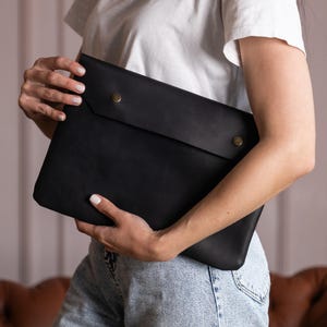 Op de afbeelding: Een zwarte leren laptoptas met een klepsluiting die wordt vastgezet met twee messingkleurige drukknopen. De tas is rechthoekig en lijkt gemaakt van glad, mat leer. Het algehele ontwerp is minimalistisch en functioneel.