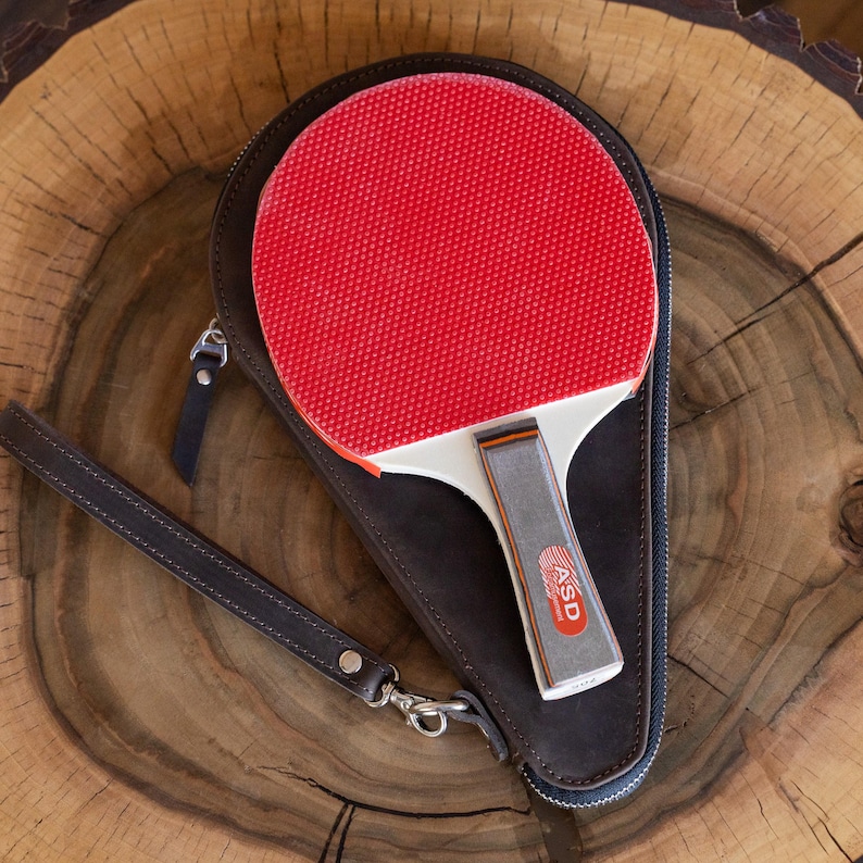 May include: A red and white ping pong paddle with a gray handle rests inside a dark brown leather case. The case has a zipper closure and a wrist strap. The paddle handle has the text "ASD".