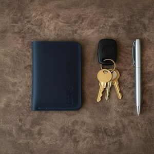 Op de afbeelding: Een marineblauwe leren portemonnee met de initialen "DWS" op de voorkant, naast een sleutelbos, een autosleutelhanger en een zilveren pen. De items zijn gerangschikt op een bruin leren oppervlak.