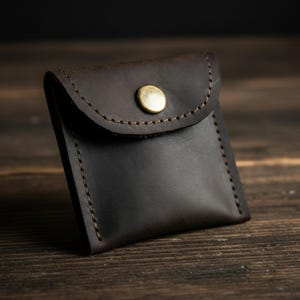 May include: A small, dark brown leather pouch with a rounded flap and a brass button closure. The edges are stitched with a contrasting thread. The pouch is square-shaped and appears to be handmade.