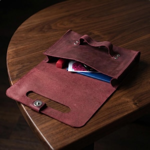 May include: A burgundy leather clutch bag with a handle and a snap closure. The open bag reveals a red lipstick, a small card with a floral image, and a blue notebook. The bag is on a dark wooden surface.