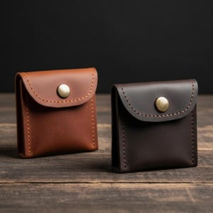 May include: Two small leather pouches in shades of brown and dark brown. Each pouch has a curved flap secured with a brass-colored snap. The pouches are stitched with a contrasting thread, and sit on a wooden surface.