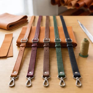 Puede incluir: Cinco correas de cuero para bolsos en tonos marrón, burdeos, marrón, verde y negro, cada una con una hebilla y un cierre plateados. Las correas están dispuestas sobre una superficie de madera con hojas de cuero y suministros de costura en el fondo.