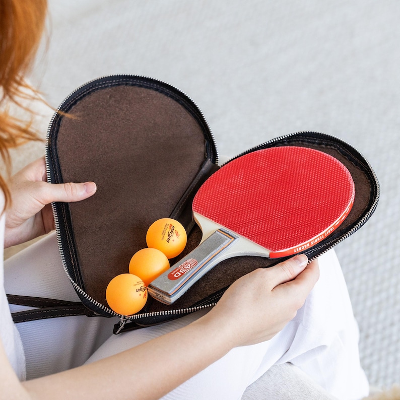 May include: A red and silver ping pong paddle rests inside a brown zippered case, alongside three orange ping pong balls. The case is open, revealing the interior. The paddle has a red rubber surface and a silver handle.