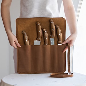 May include: A brown leather knife roll with five knives. The knives have wooden handles and silver blades. The roll is open, displaying the knives in individual slots. A brown leather strap is visible.