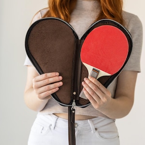 May include: A heart-shaped brown leather ping pong bat case with a zip, holding a red and white ping pong bat. The case has a wrist strap for easy carrying.