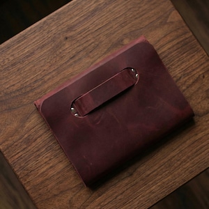 May include: A burgundy leather clutch bag with a handle. The bag is rectangular with a handle secured by silver rivets. The leather has a textured appearance and a rich, deep colour.
