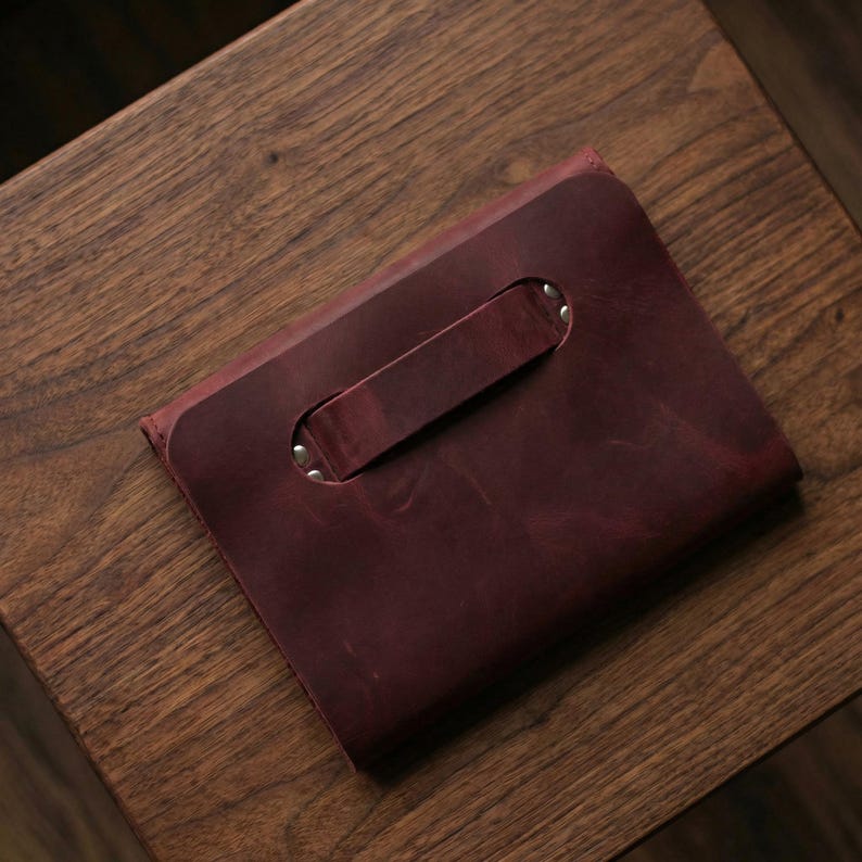May include: A burgundy leather clutch bag with a handle. The bag is rectangular with a handle secured by silver rivets. The leather has a textured appearance and a rich, deep colour.