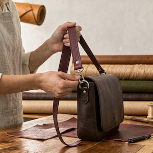 May include: A dark brown leather crossbody bag with a burgundy strap. The bag has a flap closure and silver-tone hardware. The strap is being attached to the bag. The bag is sitting on a wooden surface.