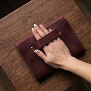 May include: A burgundy leather clutch bag with a handle, secured with metal rivets. The rectangular bag is held by a hand, resting on a wooden surface. The bag has a simple, classic design.