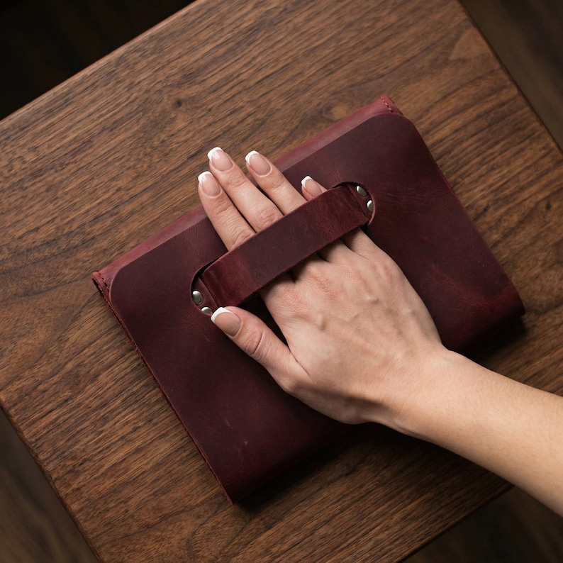 May include: A burgundy leather clutch bag with a handle, secured with metal rivets. The rectangular bag is held by a hand, resting on a wooden surface. The bag has a simple, classic design.