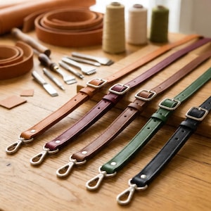 May include: A collection of leather straps in various colors, including brown, burgundy, green, and black, with silver-tone metal hardware. The straps are arranged on a wooden surface, with leather crafting tools and spools of thread in the background.