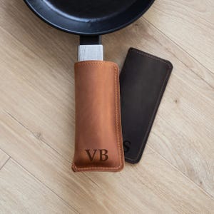 May include: A close-up shot of a black frying pan with a light brown leather handle cover and a dark brown leather handle cover. The light brown cover has the initials "VB" engraved on it. The handle covers are designed to protect hands from heat.