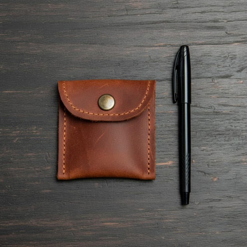 May include: A small, brown leather pouch with a button closure and stitched edges. A black pen is positioned next to the pouch on a dark wooden surface. The pouch is square with a rounded flap.