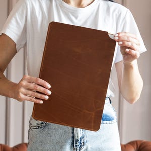 Könnte beinhalten: Eine braune Tablet-Hülle aus Leder mit einer genähten Bordüre. Die rechteckige Hülle hat oben einen Ausschnitt. Das Leder hat ein strukturiertes Aussehen und eine warme, natürliche Farbe.