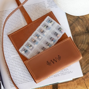 Può includere: Un astuccio per pillole in pelle marrone con un organizer per pillole in plastica trasparente all'interno. L'organizer è etichettato con i giorni della settimana e am/pm. L'astuccio ha un cinturino e una "W" decorativa con foglie.