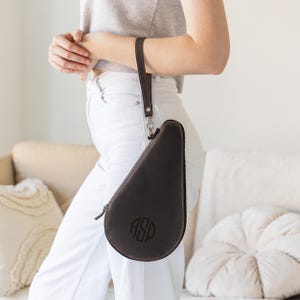 May include: Dark brown leather case with a zipper closure and a wrist strap. The case has a teardrop shape and features a monogram with the initials "ASP" in a circular design. The case is held by a person wearing a gray top and white pants.