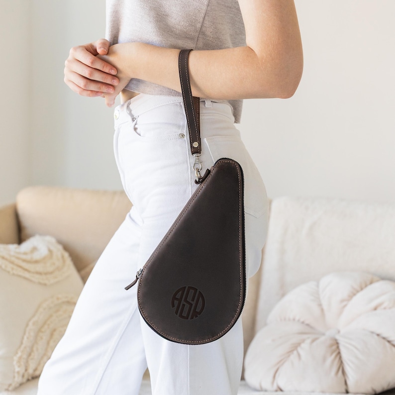 May include: Dark brown leather case with a zipper closure and a wrist strap. The case has a teardrop shape and features a monogram with the initials "ASP" in a circular design. The case is held by a person wearing a gray top and white pants.
