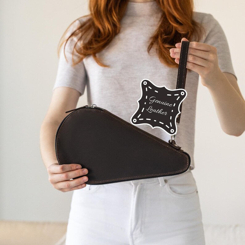 May include: A dark brown leather case with a zipper closure and a matching handle. The case has a unique teardrop shape. A tag that says "Genuine Leather" is attached to the handle.