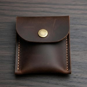 May include: A small, dark brown leather pouch with a rounded flap and a brass snap closure. The edges are stitched with light-colored thread. The pouch is likely designed to hold small items like coins or cards.