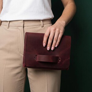 May include: A burgundy leather clutch bag with a handle. The rectangular bag has a flap closure and a strap handle on the front. The bag is held in front of a person wearing a white top and beige trousers.