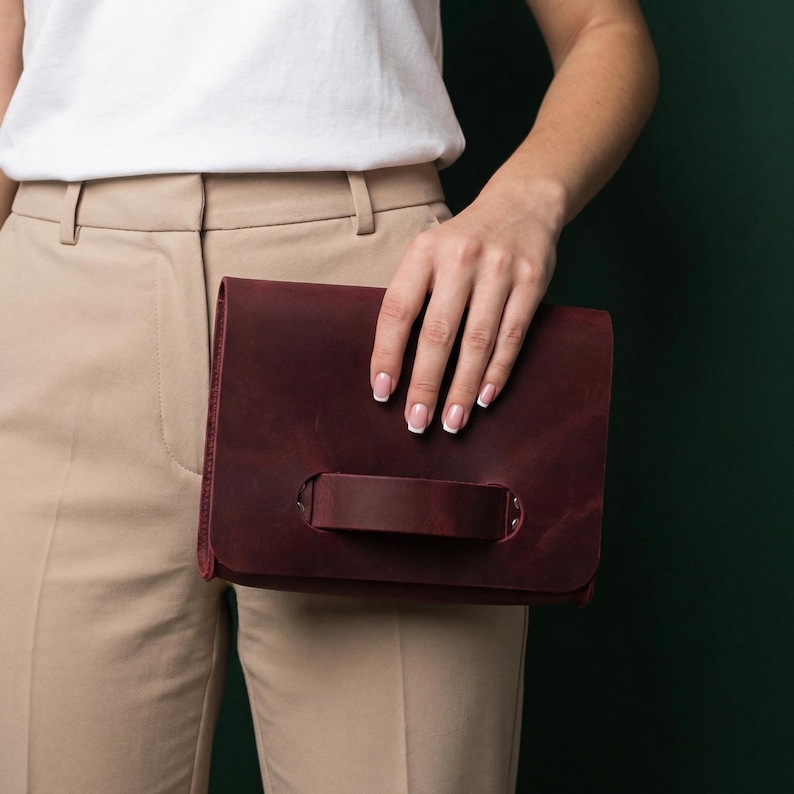 May include: A burgundy leather clutch bag with a handle. The rectangular bag has a flap closure and a strap handle on the front. The bag is held in front of a person wearing a white top and beige trousers.