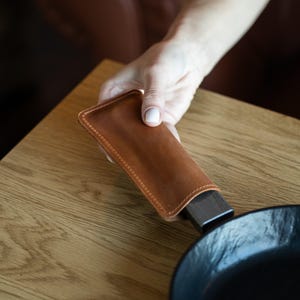 May include: A brown leather skillet handle cover with stitched edges. The cover is being held over a black cast iron skillet. The handle cover is designed to protect hands from heat when handling a hot skillet.