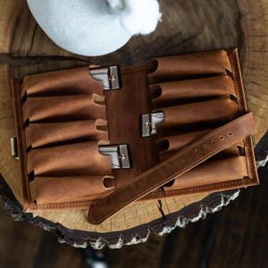Può includere: Astuccio arrotolabile in pelle marrone con più scomparti, assicurato da chiusure metalliche. È visibile un cinturino in pelle abbinato con fibbia. L'astuccio è progettato per contenere oggetti ed è esposto su una superficie di legno.