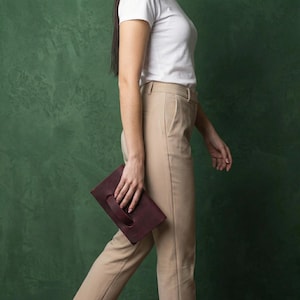 May include: A burgundy leather clutch with a handle is held by a person walking. The clutch is rectangular and appears to be made of smooth leather. The person is wearing a white t-shirt and beige trousers.