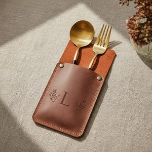 May include: A brown leather utensil holder with a gold spoon and fork. The holder has the letter "L" and leaf designs. The spoon and fork are gold with black handles. The holder is on a brown leather placemat.