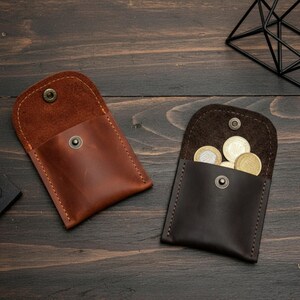 May include: Two leather coin purses, one brown and one dark brown, are displayed on a wooden surface. The brown purse is closed, while the dark brown purse is open and filled with coins. Both purses have a snap closure.