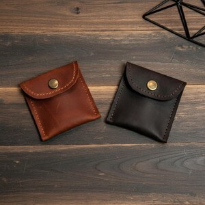 May include: Two small, square leather pouches in shades of brown and dark brown. Each pouch has a flap closure secured by a brass button. The pouches are stitched with contrasting thread, suggesting handmade craftsmanship.